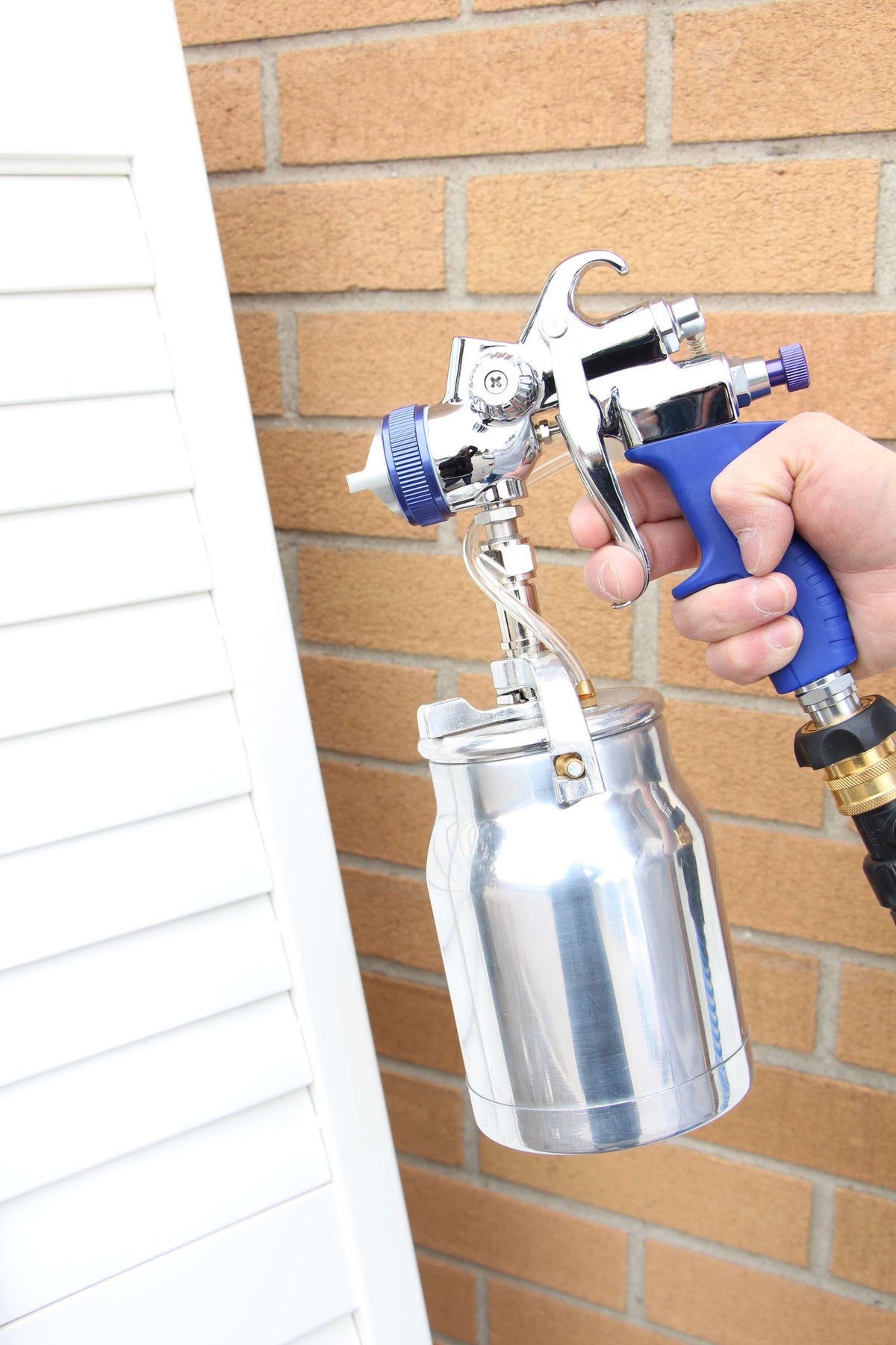Fuji Spray T70 bottom-feed spray gun spraying a white louvered shutter beside a brick wall.